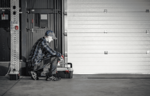 A person in a plaid shirt and cap kneels beside a toolbox in a garage, surrounded by gray walls and a large garage door.