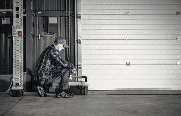 A person in a plaid shirt and cap kneels beside a toolbox in a garage, surrounded by gray walls and a large garage door.