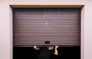 A man peeks through a partially opened brown garage door, smiling against a light pink wall backdrop.
