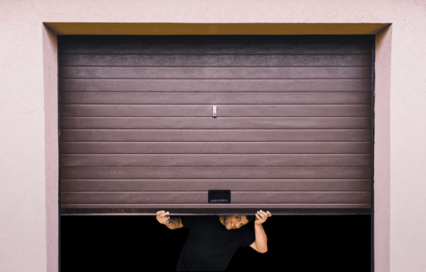 A man peeks through a partially opened brown garage door, smiling against a light pink wall backdrop.