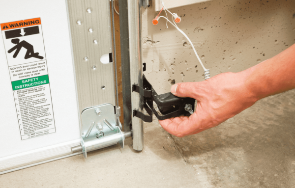 A hand adjusts a safety sensor on a garage door track, near a warning sign with safety instructions and a list of precautions.