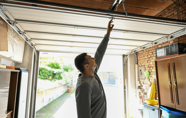 A person reaches up to a garage door opener while standing inside a bright garage filled with storage items and greenery outside.