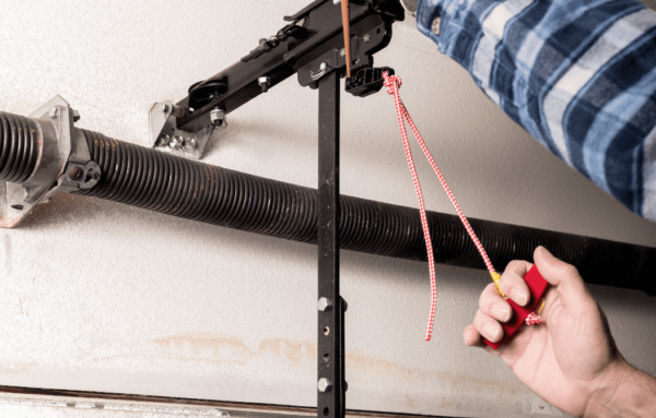 A person adjusts a garage door spring mechanism, holding a red and white rope while inspecting the metal components above.