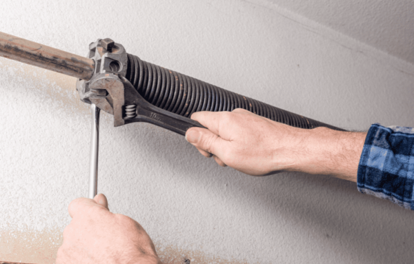 A person adjusts a garage door tension spring using a wrench and an Allen key, positioned against a wall.