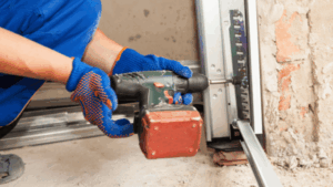 A close-up view of a technician's hands wearing blue and orange work gloves, using a cordless drill to fasten a roller or component into the metal vertical track of a garage door.