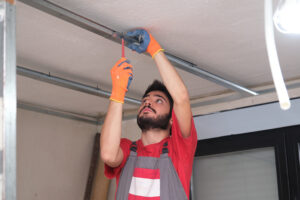 Construction worker wearing a red shirt, grey overalls, and orange and blue gloves, looking up while using a screwdriver to attach a metal stud or track to the ceiling, assembling a frame for a suspended or drop ceiling.