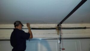 A man wearing a cap and dark shirt is working on a garage door spring, reaching up to adjust or repair it.